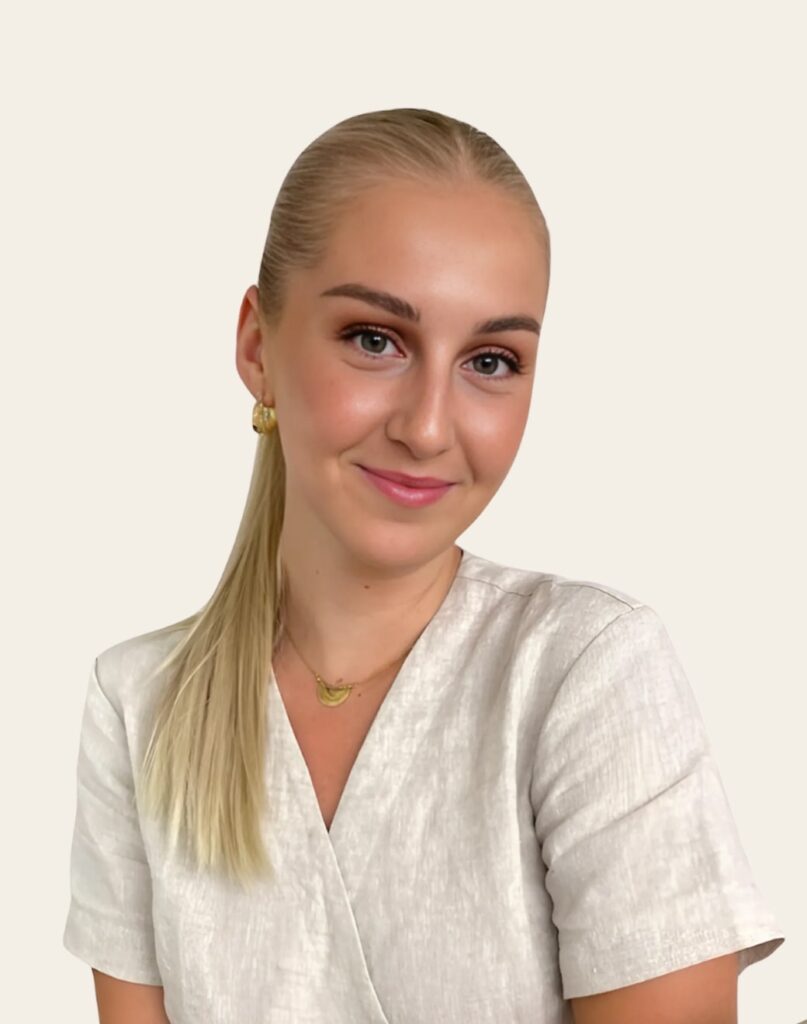 A woman with long, straight blonde hair tied back, wearing a light-colored top, poses with a slight smile against a beige background. She accessorizes with hoop earrings and a delicate necklace.
