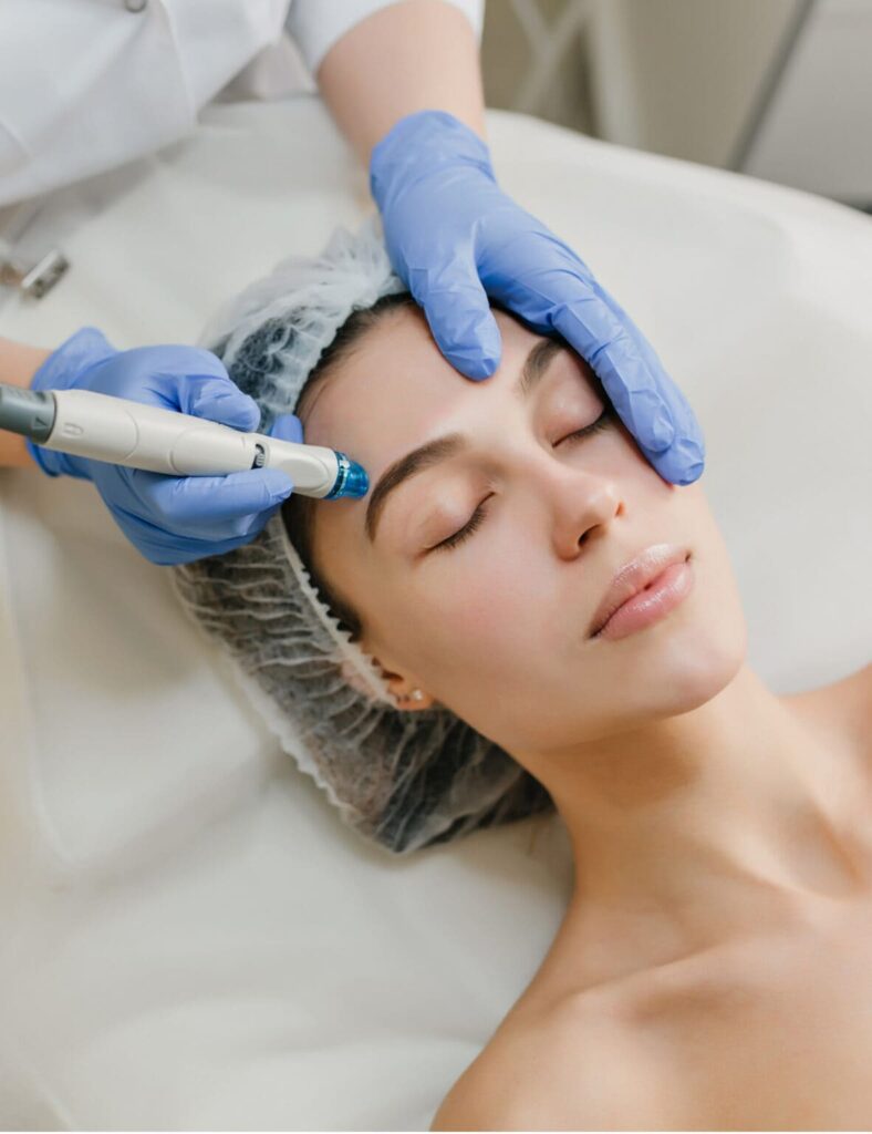 A woman with a hairnet lying on a treatment table while receiving a facial treatment. A person wearing blue gloves is using a handheld device on her skin.