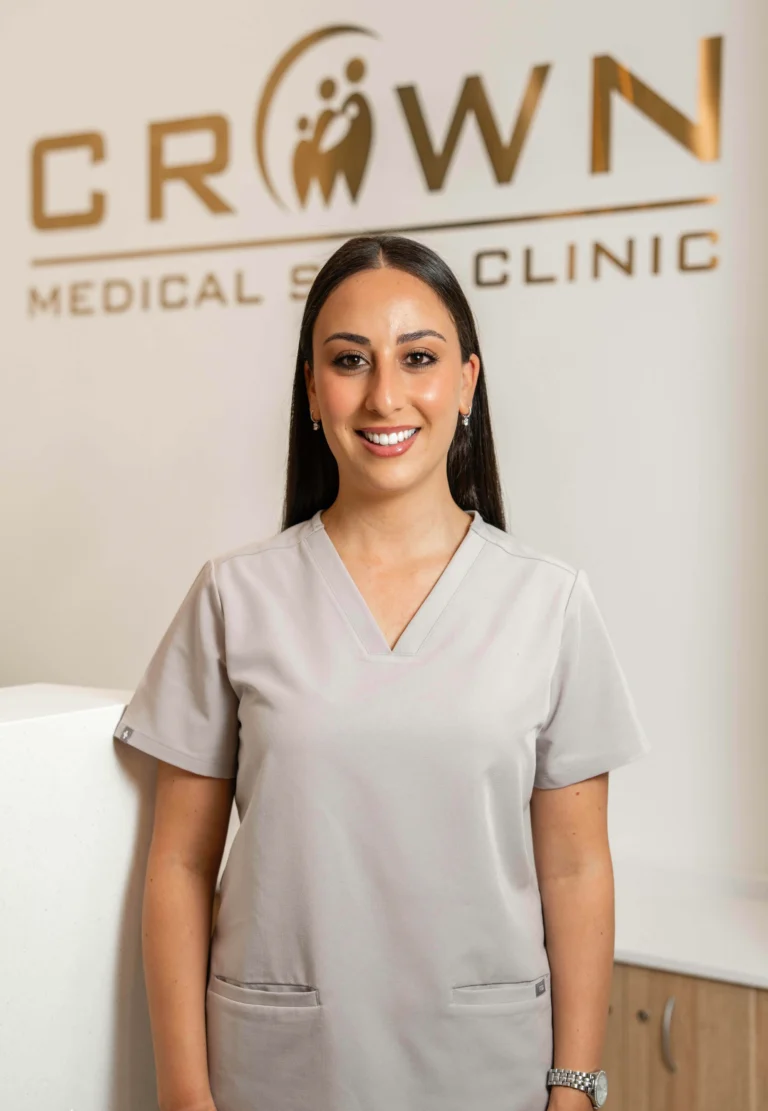 A female medical professional wearing a light grey uniform and standing in front of a sign that reads 'CROWN MEDICAL CLINIC'. She is smiling and has long dark hair.