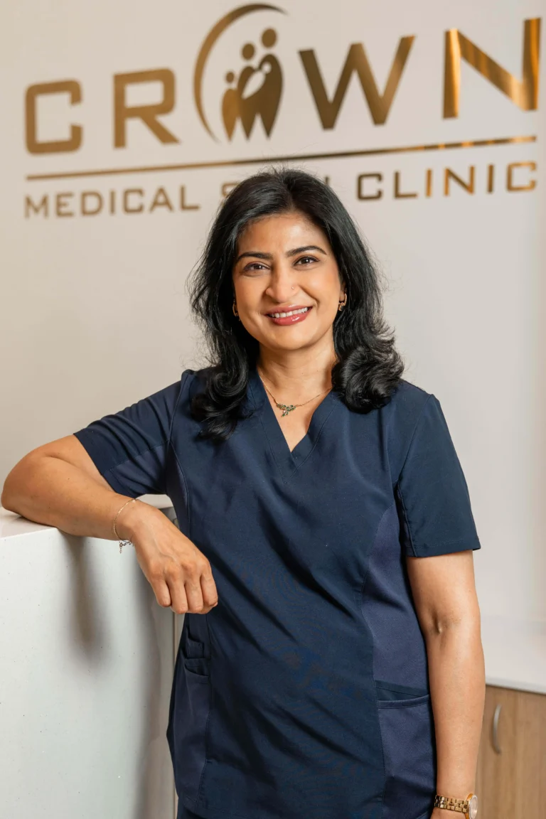 A woman in medical scrubs smiles while leaning against a reception desk. A sign in the background reads "Crown Medical Skin Clinic." The setting appears to be a clinic or medical office.