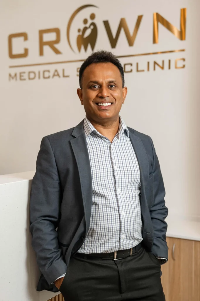 A man in a gray blazer and checkered shirt smiles while standing against a wall with the words 'CROWN MEDICAL CLINIC' in gold letters. The setting appears to be a modern medical office.